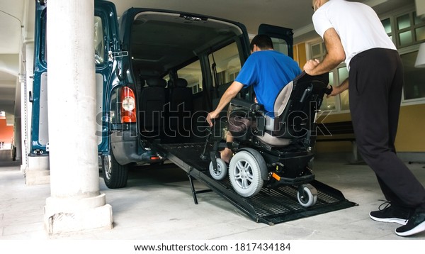 Man on wheelchair using accessible\
vehicle with ramp for transportation with driver\
helping.