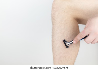 A Man On A White Background Shaves His Legs. Hairy Legs And Care For Them. Gender Equality Concept. A Close-up Showing The Foam, Razor, And Shaved Hair On A Smooth Leg.