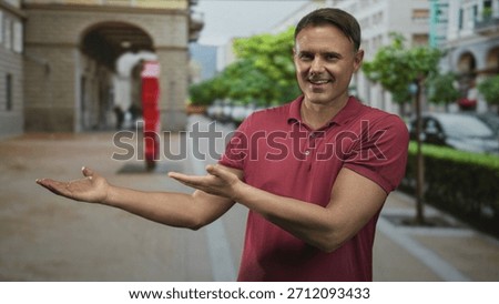 Man with open palms gesturing to a red sculpture and arch on a city street, wearing red polo; friendly invitation.