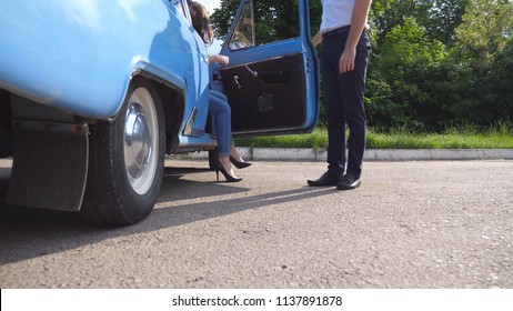 Man Opens And Holds The Door Of Old Car For The Beautiful Young Woman In High Heels Shoes. Guy Opening Door Of Vintage Automobile For Female Passenger. Girl Getting Into Retro Auto. Slow Motion.