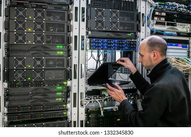 A Man Opens A Laptop Near The Racks With Computer Equipment. The System Administrator Works In The Server Room. The Programmer Makes A Backup Copy Of The Data Center Database.