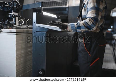Man operating controls of a large offset printing press, with stacks of paper ready for production in a modern factory setting