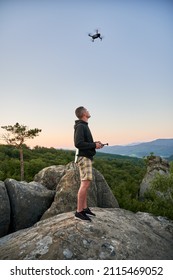 Man operating drone using remote controller. Man using drone at sunset for photos and video making while standing on top of high boulder in the mountains.