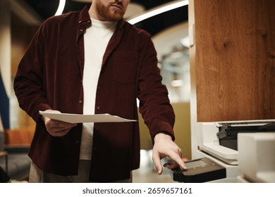 Man operating printer while holding documents in modern coworking office setting, focusing on using device for copying or scanning paperwork, face partially visible - Powered by Shutterstock - Get 15% off with code: PIKWIZARD15