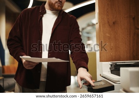 Man operating printer while holding documents in modern coworking office setting, focusing on using device for copying or scanning paperwork, face partially visible