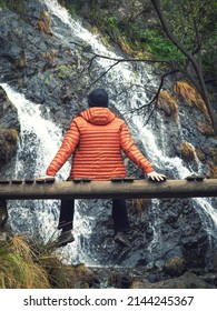 Man in orange down jacket sitting on a wooden bridge admiring a waterfall in the middle of nature. Tranquility, peace and freshness.