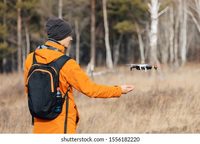 A man in an orange jacket, with a backpack and a hat, controls a quadcopter drone outdoors in autumn. The guy holds out his hand to the drone.