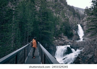 A man in an orange jacket and backpack is standing in the middle of a bridge looking at a waterfall. Concept of adventure and exploration. The man is hiking through nature.