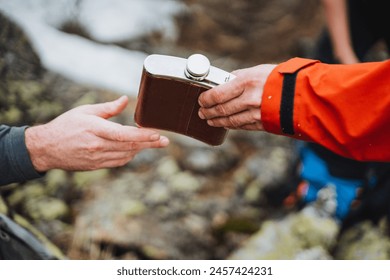 A man in an orange jacket is making a gesture with his thumb and finger, handing a flask to another man during a recreational event - Powered by Shutterstock