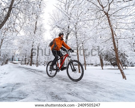 A man in an orange jacket rides a bicycle in a winter park among snow-covered trees. Active lifestyle in winter in the city