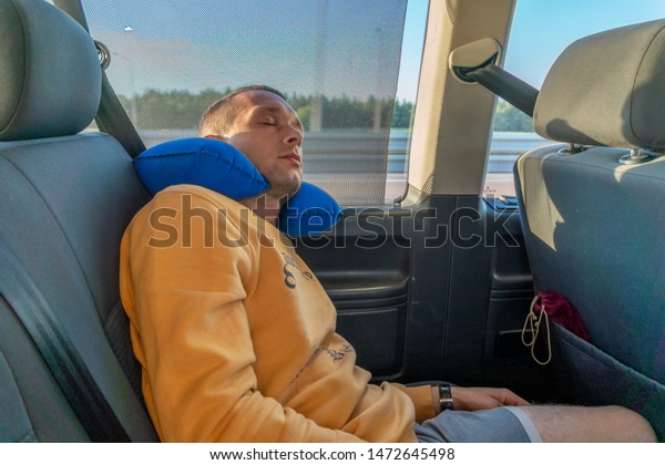 A man in an orange jacket sleeps in a car with a\
blue pillow on the road.