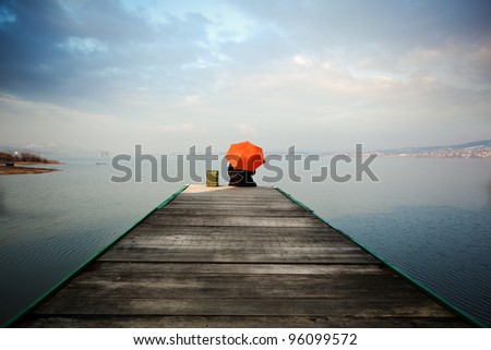 The man with the orange umbrella is sitting on a fishing pier, thinks, and he's looking at the blue lake