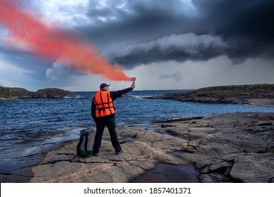 Man In An Orange Vest Gives A Signal With A Smoke Bomb. Person Brandishes A Red Smoke Bomb. SOS Signal. Man With A Torch Against The Sky With Gray Clouds. Man Is In Distress On A Rocky Island.