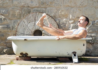 Man in the outdoor bathtub washes his leg with brush. Hygiene in the tub in the yard.