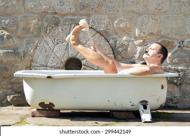 Man in the outdoor bathtub washes his leg. Mustachioed man washes his feet with a brush in the outdoor tub. Hygiene in the outdoor bathroom. Thorough hygiene with scrub brush in the bath.
