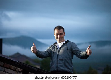 Man Outdoors on the Roof of the Building Making Success Gesture with Cloudy Hills on Background