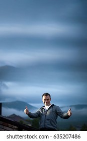 Man Outdoors on the Roof of the Building Making Success Gesture with Cloudy Hills on Background