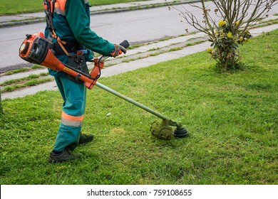 Man in overall and safety helmet trims overgrown grass by grass cutter