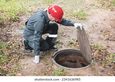 a man in overalls opened a sewer hatch and looks into a septic tank. Cleaning of sewers and drains.