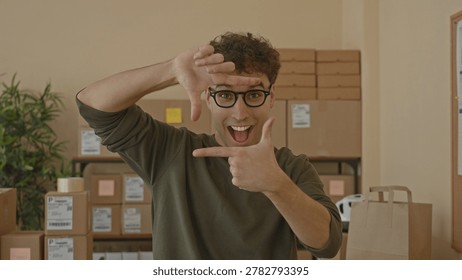 Man with packages frames his face with hands and points at camera while smiling amid stacked parcels on shelves inside a building; excitement.