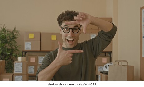 Man with packages frames his face with hands and points at camera while smiling amid stacked parcels on shelves inside a building; excitement.
