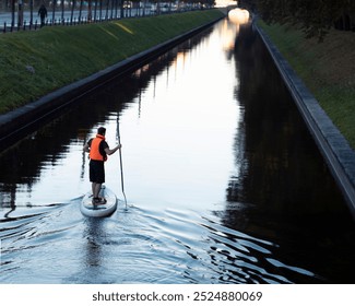 Man paddleboarding on calm canal water in city. Side view of paddler wearing life vest during sunset. - Powered by Shutterstock