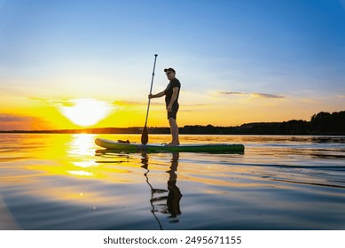 Man Paddleboarding at Sunset on a Lake. A man stands on a paddleboard in a lake as the sun sets, casting a golden glow on the water. - Powered by Shutterstock