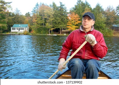 A Man Paddles A Canoe On A Lake In Vermont During Fall Foliage Season. Summer Cabins Line The Shore In The Background.