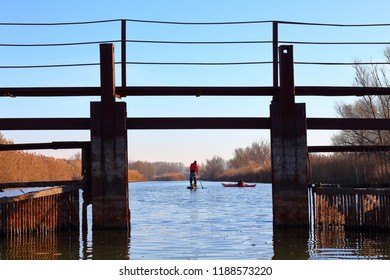 Man Paddles A Red Kayak And Man Standing With A Paddle On The Stand Up Paddle Board (paddleboard, SUP) On The River Or Lake Near Old Rusty Bridge In Fall Season. Autumn And Winter Kayaking