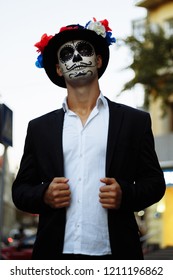 a man with a painted face of a skeleton, a dead zombie, in the city during the day. day of all souls, day of the dead, halloween, ghost walk in a hat