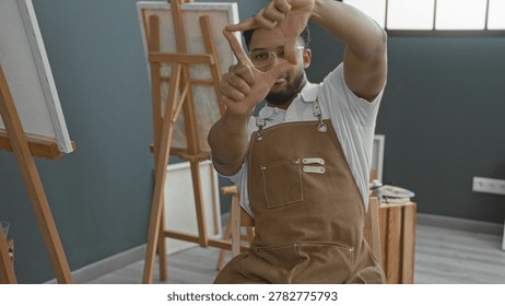 Man painter in apron frames scene with hands making a viewfinder in studio; contemplative creativity.