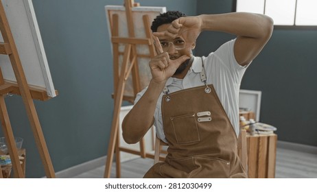 Man painter in apron frames scene with hands making a viewfinder in studio; contemplative creativity.