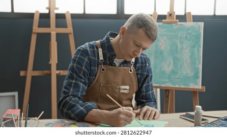 Man painter in apron holds smartphone and brush, paints at easel while rubbing forehead in studio; creative frustration.