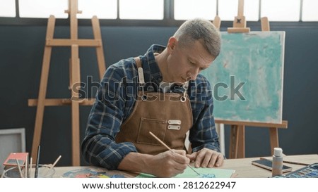 Man painter in apron holds smartphone and brush, paints at easel while rubbing forehead in studio; creative frustration.