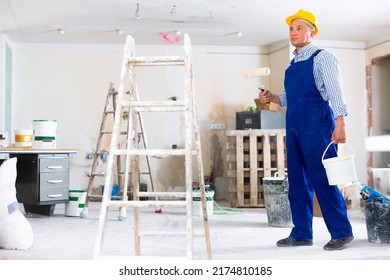 Man Painter In Coverall And Hardhat Carrying Bucket And Paint Roller. Male Builder Working In Construction Site.