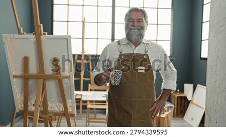 Man painter drinking from mug in studio, wearing apron and beard, standing by easel and canvas near large window; contentment creativity.
