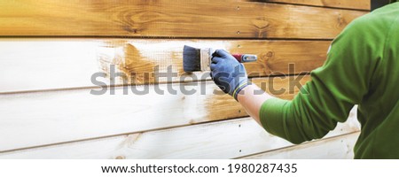man painting wood house exterior siding with brown protective paint. banner copy space