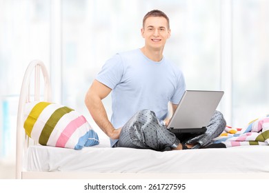 Man In Pajamas Working On A Laptop Seated On A Bed At Home