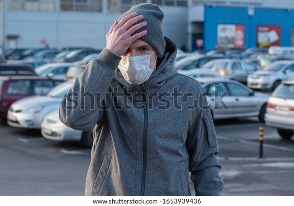 man in the\
parking lot. Feels unwell, headache.  Holding his hand to his head.\
 On the face is a white medical mask.  Protection against bacteria,\
viruses.  Symptom of coronavirus.\
