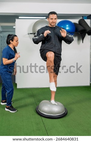 Man performing balancing exercise on a bosu ball with support from a female physiotherapist in a rehabilitation center