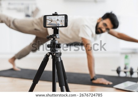 Man performing yoga pose while recording content for a fitness blog using smartphone setup on tripod. Promotes exercise, mindfulness, and home health routines for an active lifestyle improvement.