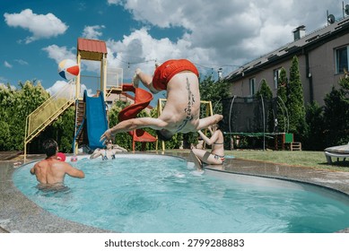 A man performs a back flip into a residential pool surrounded by friends and a water slide. Happy group enjoys summer play and splashing in the backyard pool.