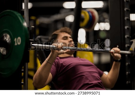 A man performs a bench press exercise in a gym, concentrating on his form as he lifts weights. The gym has various equipment and colorful plates on the barbell.
