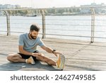 A man performs a seated hamstring stretch on a wooden boardwalk by the water, with sailboats and a city skyline in the background. 

