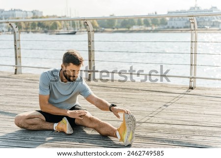 A man performs a seated hamstring stretch on a wooden boardwalk by the water, with sailboats and a city skyline in the background. 

