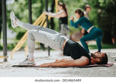 A man performs a single-leg glute bridge on a mat as part of an outdoor workout. He trains with others in a park setting during a group fitness session.