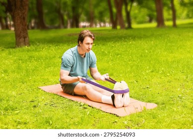 A man performs a stretching exercise with a resistance band on a fitness mat at a park, focusing on his outdoor athlete workout. This high-quality photo shows resistance band stretching, effective resistance band training for flexibility and mobility, and outdoor resistance band workout as part of a fitness routine in a park setting.