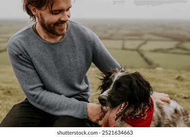 A man with a pet, wearing a blue sweater, sits in nature outdoors with his dog. The man and his dog pet, wearing a red bandana, enjoy a moment in nature. Man and dog pet bonding in nature landscape. - Powered by Shutterstock