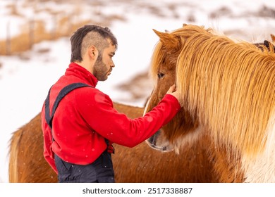 A man is petting a horse with a long mane. The man is wearing a red shirt and blue overalls. The horse is brown and white. Horse in the cold snowy winter of Iceland - Powered by Shutterstock