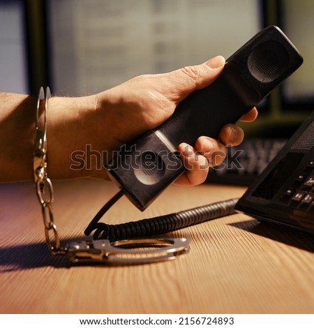 A man at the phone in handcuffs on his hands, close-up. Office desk in a dark night room with computer monitors